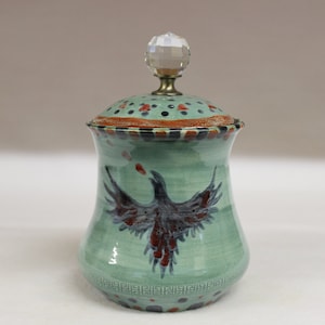 May include: A ceramic jar with a lid, featuring a hand-painted design of a bird with outstretched wings. The jar is light green with a decorative rim and a crystal knob on top. The bird is dark gray and red.
