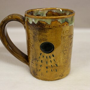 May include: Handmade ceramic mug with a unique design. The mug features a golden-brown exterior with etched hieroglyphs and a black circular accent. The rim has a green and brown glaze. The handle is a matching golden-brown color.