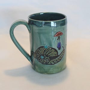 May include: A teal ceramic mug with a curved handle. The mug features a hand-painted design of a snail and a mushroom. The snail has a detailed shell and is decorated with teal and brown colors. The mushroom is red and purple.
