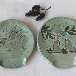 May include: Two round ceramic spoon rests with a light green glaze and dark green polka dots. One features two mice interacting with a dragonfly, while the other shows three mice perched on a branch with blue berries. Small dark brown pinecones sit above.