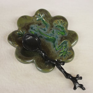 May include: A ceramic spoon rest with a frog design and a black spoon. The spoon rest is flower-shaped with a green and blue glaze, featuring a frog illustration. The black spoon has a branch-like handle.