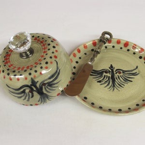 May include: A ceramic butter dish with a matching plate and butter knife. The dish has a crystal knob and is decorated with red and black dots and a black bird design. The plate and knife also feature the bird design.