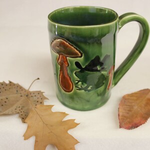 May include: A green ceramic mug with a whimsical design featuring a mushroom and a frog. The mug has a large handle and is decorated with brown and black accents. The mug is approximately 4 inches tall.