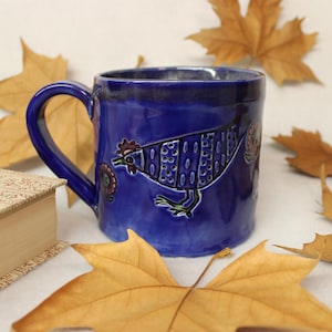 May include: A cobalt blue ceramic mug with a hand-painted rooster design. The mug has a curved handle and is surrounded by autumn leaves and a book. The rooster is depicted in white, black, and red.