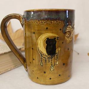 May include: A ceramic mug with a brown and gold glaze, featuring a black owl silhouette on a crescent moon. The mug has a dark brown handle and is decorated with small black dots and celestial designs. The rim has a dark blue-green glaze.