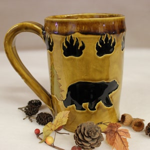 May include: A ceramic mug with a brown and tan glaze, featuring a black bear silhouette and bear paw prints. The mug has a large handle and is decorated with a forest theme. The mug is surrounded by decorative fall foliage.