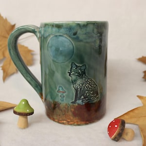May include: A ceramic mug with a teal glaze featuring a fox silhouette under a full moon. The mug has a dark green handle and a brown base. Decorative wooden mushrooms and fall leaves surround the mug.