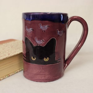 May include: A ceramic mug with a burgundy base and a dark blue rim. The mug features a black cat design with yellow eyes and small bird illustrations. The handle is burgundy, and a book is visible in the background.