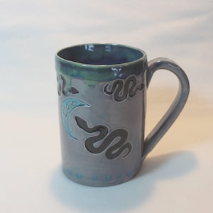 May include: A ceramic mug with a gray and teal glaze. The mug features a handle and is decorated with a crescent moon and snake design in black and teal. The rim is a darker teal color.