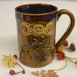 May include: A brown ceramic mug with a handle, featuring a raised design of a rabbit, crescent moons, and stars. The mug has a dark green interior and is decorated with a glossy finish. The mug is approximately 5 inches tall.