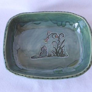 May include: A rectangular ceramic dish with rounded corners and a glossy sage green glaze. The rim features a dark dotted pattern. The center of the dish displays a debossed illustration of a small creature sitting beside a tall, drooping flower.
