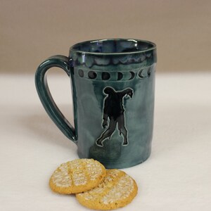 May include: A teal ceramic mug with a black zombie graphic and moon phase design. The mug has a large handle and is paired with two round cookies with white icing.