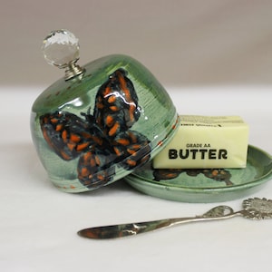 May include: A green ceramic butter dish with a butterfly design in orange and black. The lid has a crystal knob. A stick of butter sits on the matching saucer, and a silver butter knife is next to it.