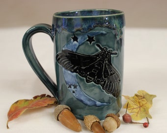 May include: A teal ceramic mug with a large handle, featuring a black moth design against a crescent moon and stars. The mug has a textured glaze and is decorated with acorns and leaves.