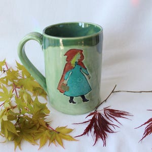 May include: A green ceramic mug with a handle, featuring an illustration of Little Red Riding Hood in a red cape and blue dress. The mug is decorated with a floral design and is surrounded by fall foliage.