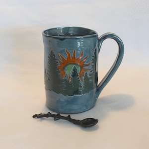May include: A teal ceramic mug with a forest and sun design. The mug features a handle and a detailed illustration of trees and a sun with orange and green accents. A small, dark spoon rests beside the mug.