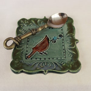 May include: A decorative spoon rest with a ceramic base and a metal spoon. The base is square with a green border and a teal center featuring a red bird illustration. The spoon has a twisted handle.