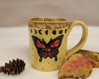May include: A ceramic mug with a yellow base, featuring a black butterfly design with red accents. The mug has a curved handle and is decorated with phases of the moon. Red dots are scattered around the butterfly. A pinecone and fall leaves are next to the mug.