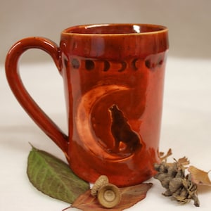 May include: A reddish-brown ceramic mug with a handle. The mug features an embossed design of a howling wolf inside a crescent moon. The mug also has embossed phases of the moon around the top. The mug is surrounded by leaves and acorns.