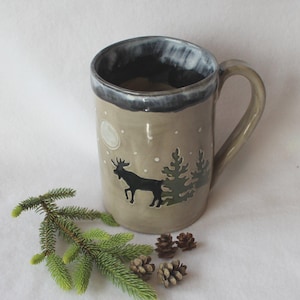 May include: A beige ceramic mug with a black moose silhouette, green trees, and a white moon design. The rim is dark blue. The mug has a curved handle. Decorative pinecones and a sprig of evergreen are in the foreground.
