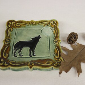May include: Square ceramic tile with a howling wolf silhouette against a light green background and a full moon. The tile is framed with a decorative brown and gold border. Decorative fall elements are in the background.