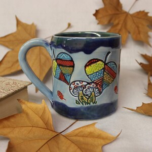 May include: A ceramic mug with a light blue handle and body, featuring colorful leaf and mushroom designs. The mug has a dark blue rim and base, with a green interior. Autumn leaves and a book are in the background.