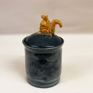 May include: A ceramic jar with a lid, featuring a golden squirrel figurine on top. The jar is dark blue with a textured finish and a decorative band near the base. A unique decorative piece for the home.