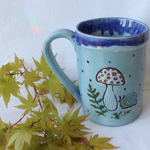 May include: A light blue ceramic mug with a whimsical mushroom and snail design. The mug has a dark blue rim and handle, with a light blue interior. The mug is decorated with a red and white mushroom, a snail, and fern. The mug is surrounded by green leaves.