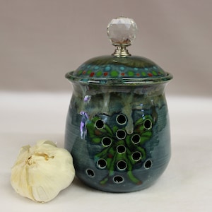 May include: A teal ceramic garlic keeper with a lid, featuring a crystal knob. The keeper has a green butterfly design with holes for ventilation. A bulb of garlic sits beside the keeper.