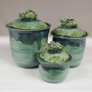 May include: Three ceramic jars in varying sizes, glazed in shades of green and blue. Each jar has a lid with a sculpted fish handle and fish-shaped designs. The jars have a textured, handmade appearance.