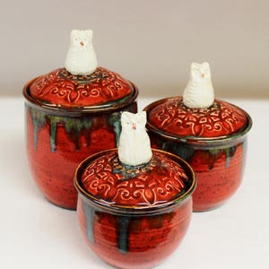 May include: Set of three ceramic jars with lids. The jars are a deep red with a textured finish and a green and blue glaze. Each lid has a white owl figurine on top. The lids have a decorative pattern.