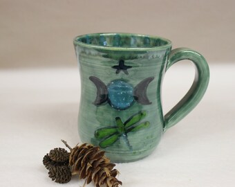 May include: Green ceramic mug with a handle, featuring a celestial design. The mug has a dragonfly, a star, and a moon phase design in black and teal. The mug is approximately 4 inches tall and has a curved shape.