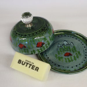 May include: A ceramic butter dish with a forest scene design. The green dish has a domed lid with a crystal knob and a matching plate. A stick of butter is next to the dish, with the word "BUTTER" printed on it.