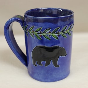 May include: A blue ceramic mug with a black bear silhouette and a green and red leaf design. The mug has a large handle and is approximately 5 inches tall. The interior of the mug is a darker blue.