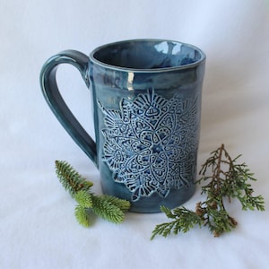 May include: A teal ceramic mug with a large, detailed embossed snowflake design. The mug has a curved handle and is surrounded by sprigs of greenery. The mug is a solid color with a glossy finish.