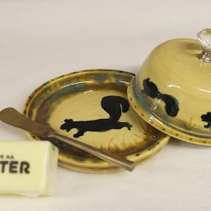 May include: A ceramic butter dish with a lid, a butter knife, and a stick of butter. The dish and lid are yellow with black squirrel silhouettes. The butter is wrapped in white paper with the word "BUTTER" printed on it.