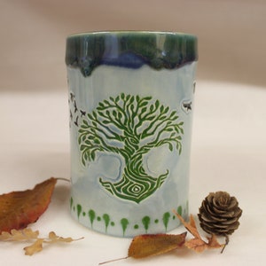 May include: A ceramic tumbler with a light blue base and a green and blue rim. The tumbler features a detailed green tree design and small bird silhouettes. The base has a row of green dots. Autumn leaves and a pine cone are next to the tumbler.