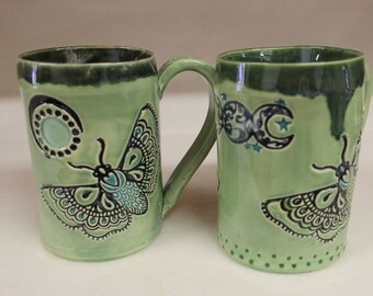 May include: Two green ceramic mugs with a shared handle. Each mug features a detailed butterfly design with teal and black accents. One mug has a moon design, the other has a crescent moon and star design.