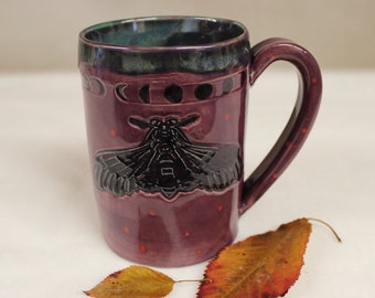 May include: A ceramic mug in shades of purple with a black moth design and phase of the moon details. The mug has a handle and is decorated with small red dots. Two autumn leaves rest near the mug.