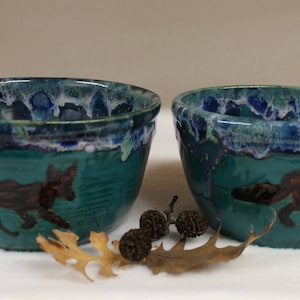 May include: Two ceramic bowls with a teal exterior and a dark blue and white rim. Each bowl features a brown fox illustration. The bowls are decorated with a leaf and small pinecones, suggesting a nature theme.