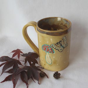 May include: A ceramic mug with a yellow-tan base color, adorned with a mushroom and butterfly design. The mug has a large handle and a brown rim. White dots are scattered across the surface. The mug is decorated with a red and blue mushroom and a butterfly.