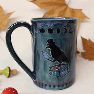 May include: A teal ceramic mug with a dark green handle. The mug features a black raven wearing a top hat and reading a book, with a crescent moon design. The mug is decorated with small dots and red and orange accents.