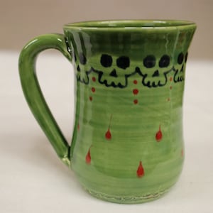 May include: Green ceramic mug with a handle, decorated with black skull designs around the rim and red teardrop accents. The mug has a glossy finish and a unique, handcrafted appearance, perfect for beverages.