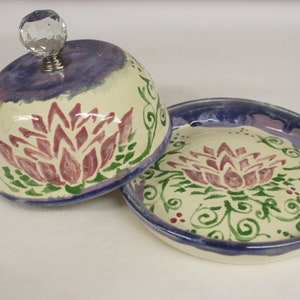 May include: A ceramic butter dish with a lid, featuring a floral design. The dish and lid are off-white with a purple and blue rim. The floral design is in shades of red and green. The lid has a crystal knob.