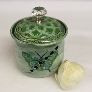 May include: A green ceramic garlic keeper with a butterfly design. The keeper has a matching lid with a crystal knob. A bulb of garlic sits next to the keeper. The keeper has a series of holes for ventilation.