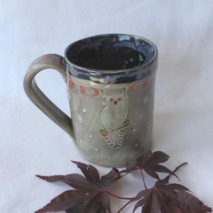 May include: A handmade ceramic mug with a gray-green glaze. The mug features an embossed owl design with red eyes, crescent moons, and stars. The rim is dark blue with a red and white trim. The handle is gray-green.