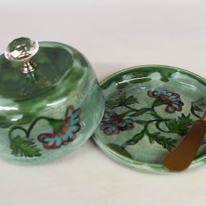 May include: A green ceramic butter dish with a matching plate and a gold-colored butter knife. The dish has a domed lid with a crystal knob. Floral designs in teal and red adorn the dish and plate.