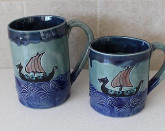 May include: Two ceramic mugs with a glossy finish, featuring a hand-painted black Viking ship with a tan sail on a dark blue, textured wave pattern. The upper portion of each mug is a lighter blue, and each has a curved handle on the right side.