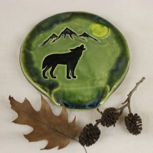 May include: A green ceramic spoon rest with a black wolf howling at the moon and mountains. The spoon rest has a dark green border and a curved shape. A brown leaf and pinecones are next to the spoon rest.
