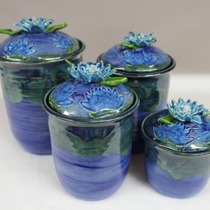 May include: Four blue ceramic jars with lids, each adorned with a blue lotus flower and green leaves. The jars vary in size, with a dark blue rim and a textured, glazed finish. The jars are a decorative home decor item.